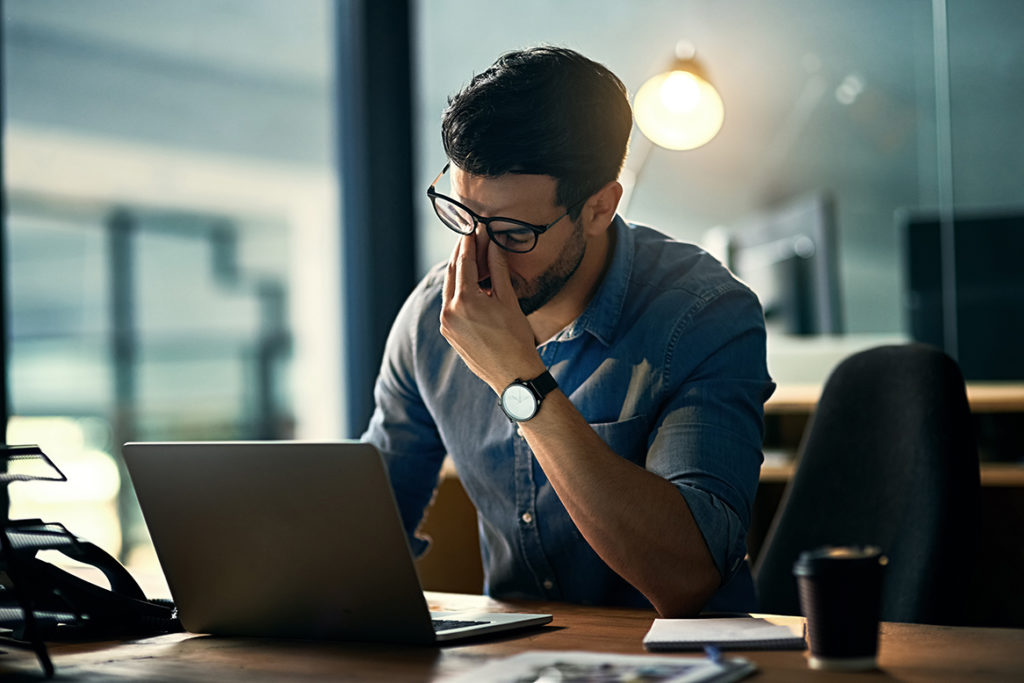 A man sitting at the computer holding the bridge of his nose in stress.